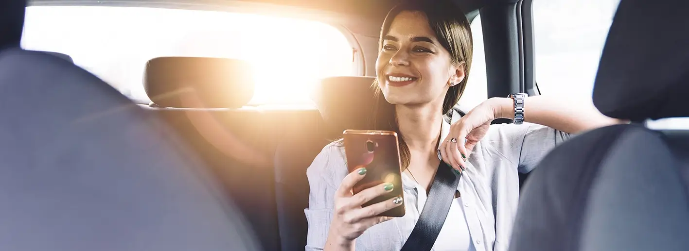 Fröhliche, positive junge Frau in Freizeitkleidung sitzt angeschnallt auf dem Rücksitz eines Autos und schaut weg, während sie ihr Handy benutzt.